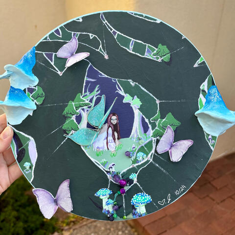 A circular, 3d, mixed media art piece. A fairy wearing a white dress crouches in the middle of a forest. She has sparkly teal wings. Purple butterflies, crystals, and blue and purple mushrooms surround her.