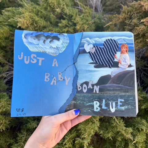 An art journal collage with blue paper, ocean paintings, a cloud painting, blue love heart shapes, and a photograph of a girl wearing blue. There is text across the spread. Text: Just a baby born blue.